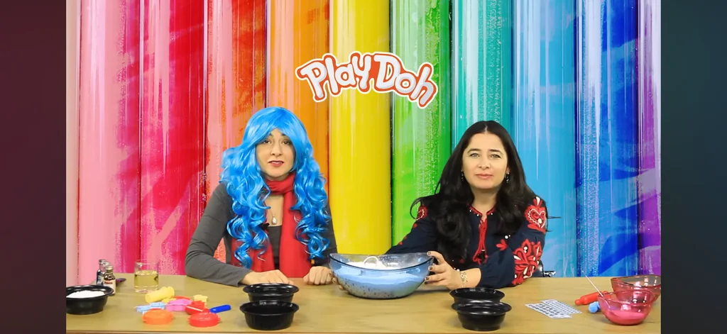 Aroub Soubh on the Waqt Al Farah YouTube series, 2015, at a table of bowls and modelling dough before a rainbow-striped wall, with a co-presenter in a blue wig.