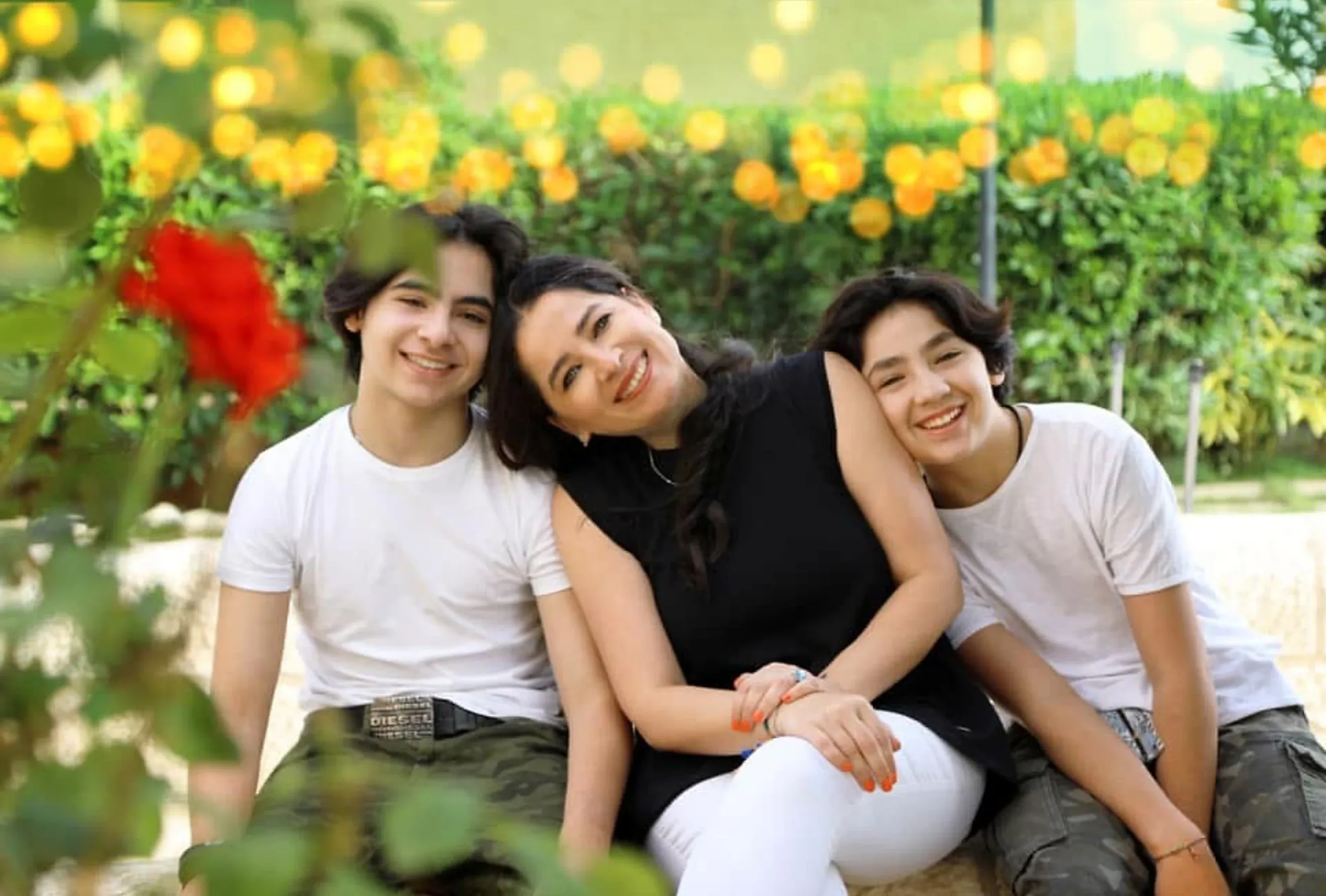 Aroub Soubh in 2019 with her sons Hisham and Tareq leaning into her from either side, a garden with a red rose and string lights behind them. Her photograph.