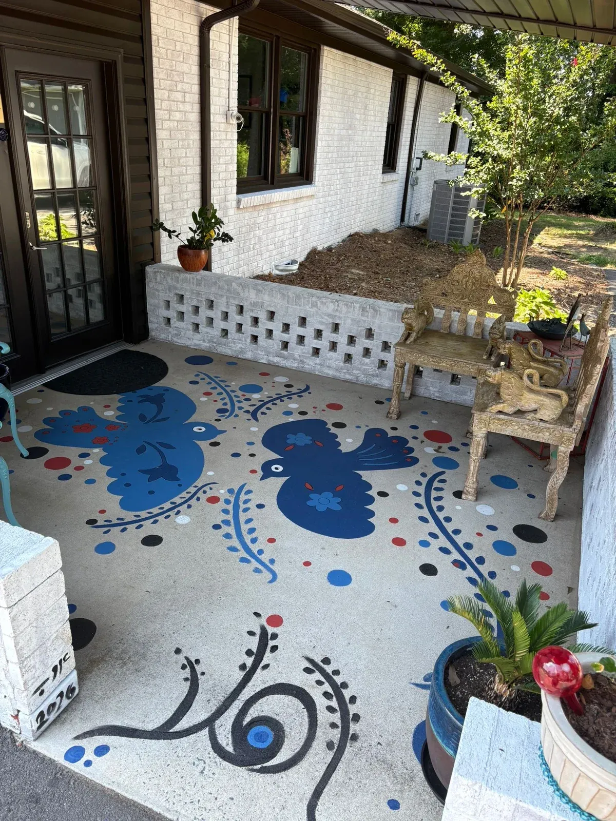 The painted floor at the entrance of her sister's salon: two cobalt birds with flowers, red, white and black dots and blue branches on pale concrete, between the glass doors and a carved bench; her signature and 2026 on the white pillar.
