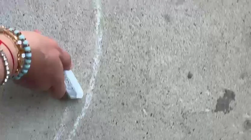 Aroub Soubh's hand, bracelets on the wrist, drawing the bird's outline in white chalk on concrete.