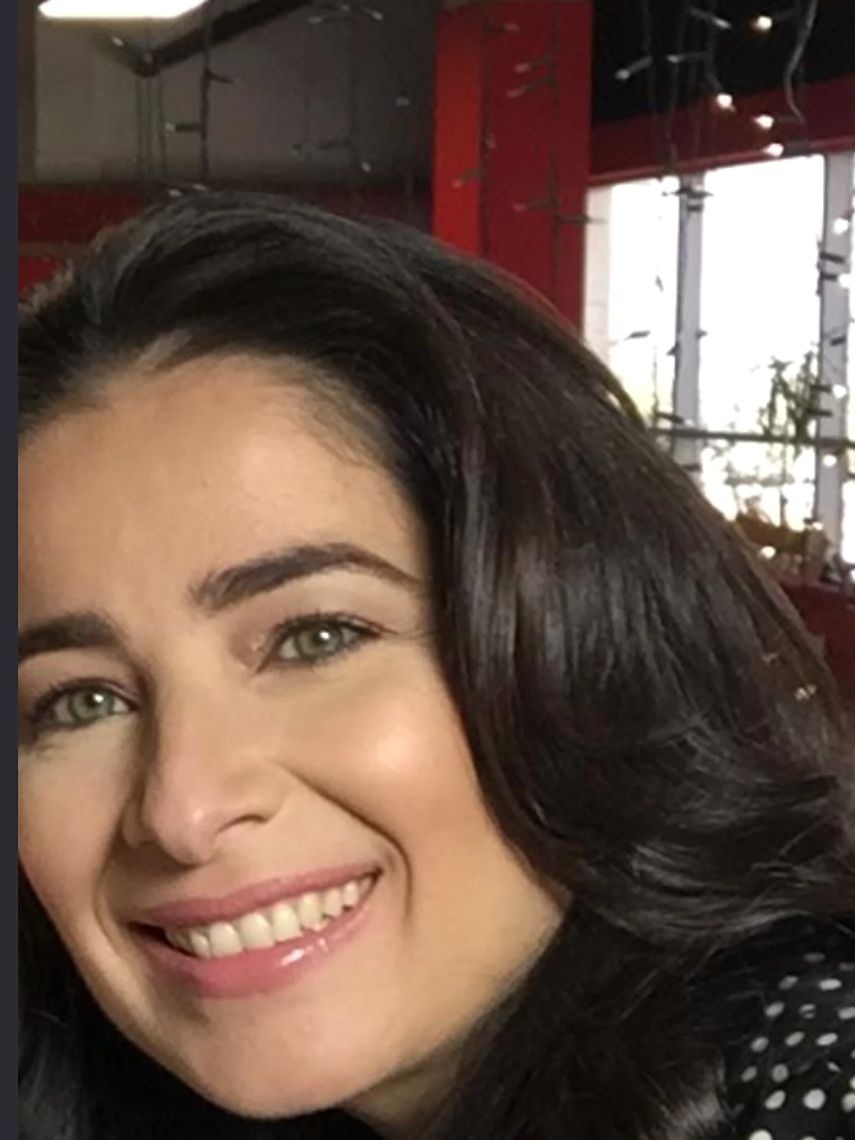 Aroub Soubh, close, smiling, in a black blouse with white polka dots, red studio walls and string lights behind her. Her photograph.