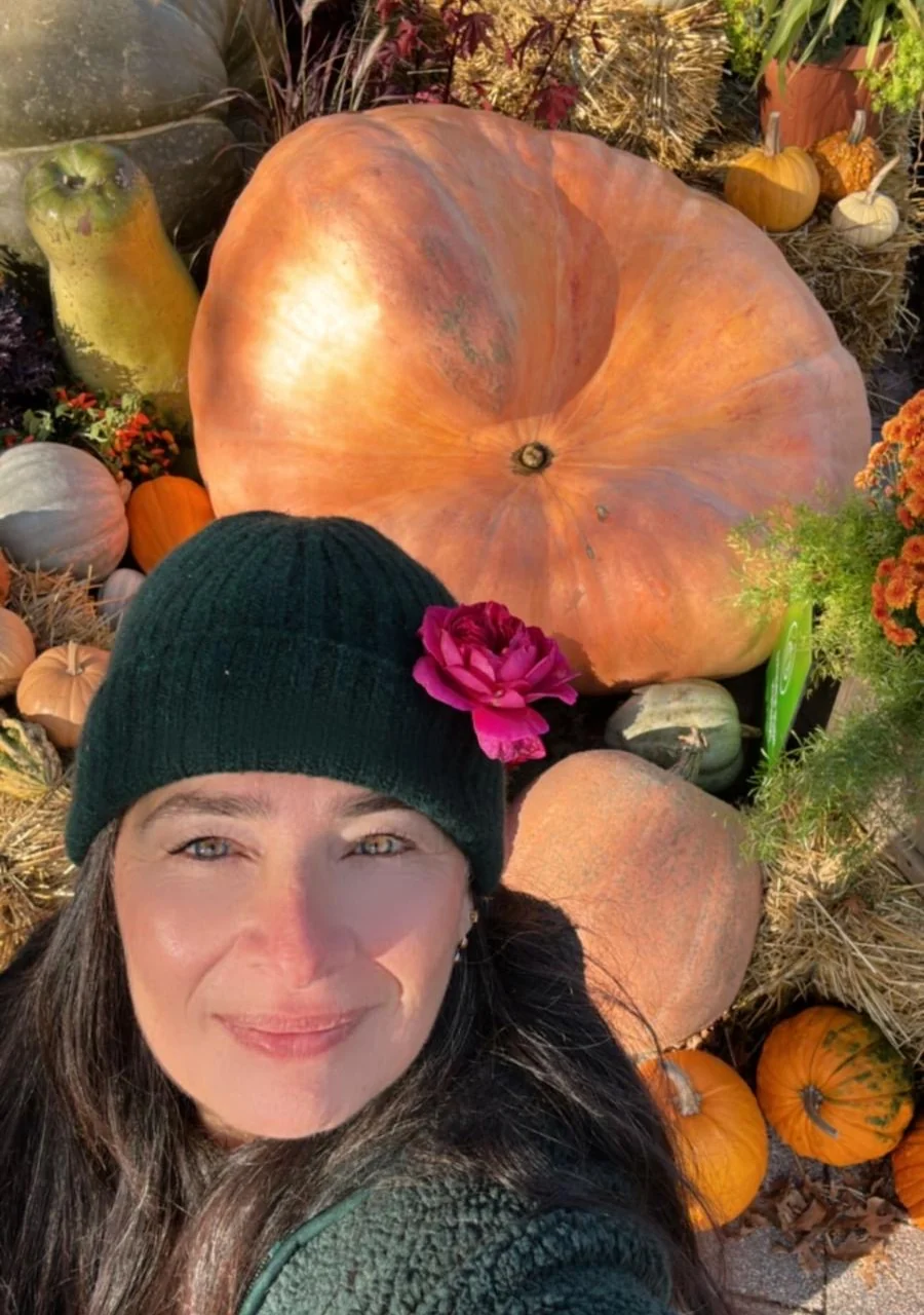 Aroub Soubh today, smiling into the camera in a green beanie with a pink rose tucked into it, a giant orange pumpkin and autumn flowers behind her, Ontario. Her photograph.